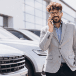 A car dealership salesman smiling on the phone in front of a row of cars