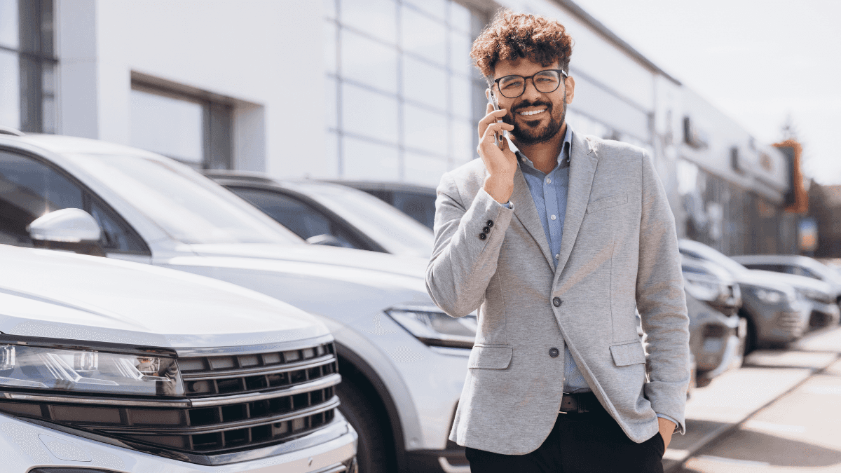 A car dealership salesman smiling on the phone in front of a row of cars