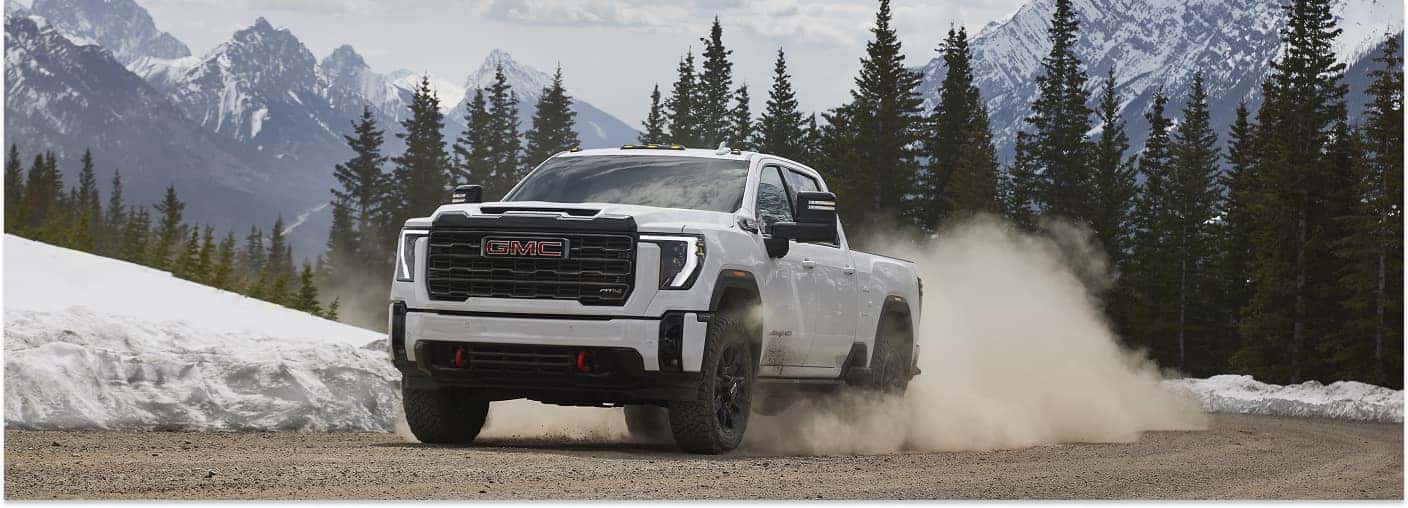 A white 2026 GMC Sierra 2500 heavy-duty pickup truck kicks up a large cloud of dust while driving along a dirt road, set against a scenic backdrop of snow-capped mountains and pine trees.