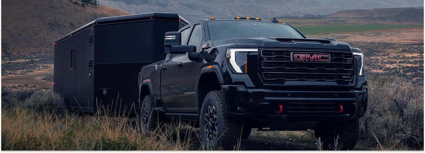 Black 2026 GMC Sierra 2500 HD towing a black enclosed trailer in a field, front three-quarter view.