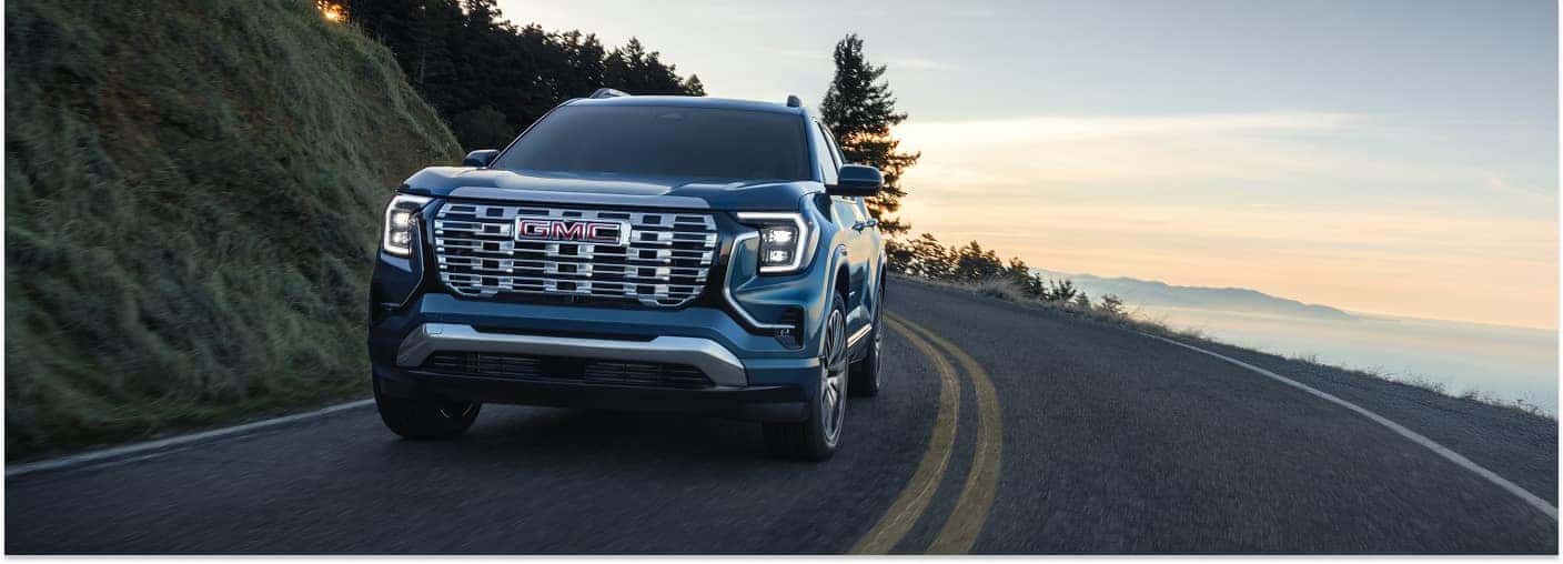 Front view of a blue 2026 GMC Terrain driving on a mountain road at sunset.