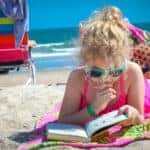 Girl in pink tank top wearing eyeglasses reading book on beach during daytime