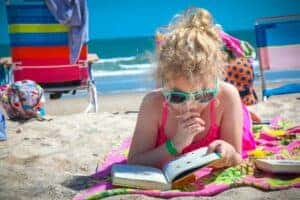 Girl in pink tank top wearing eyeglasses reading book on beach during daytime