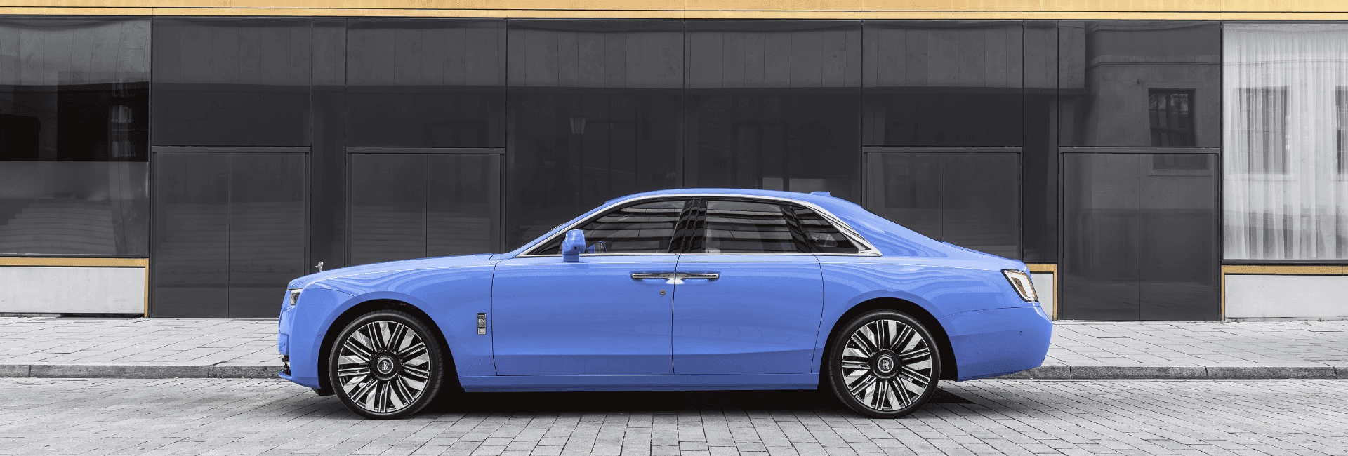Light blue Rolls-Royce Ghost shown in full side profile, parked on a paved street in front of a modern building with large dark glass windows, highlighting the vehicle’s long, elegant body, chrome accents, and multi-spoke wheels.