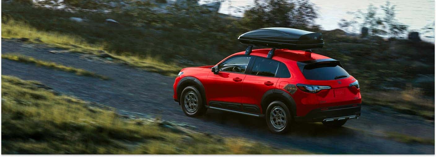 Rear three-quarter view of a red 2026 Honda HR-V driving on a gravel road with a roof cargo box.
