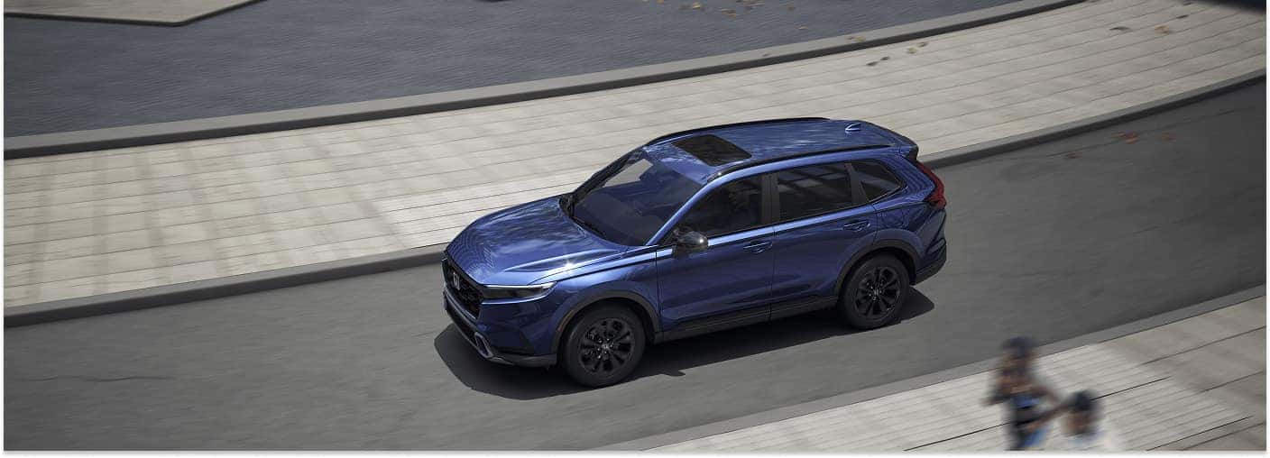 High-angle front three-quarter view of a blue 2026 Honda CR-V Hybrid driving on an urban street.