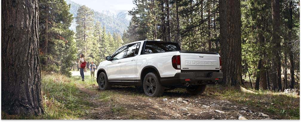 White 2026 Honda Ridgeline pickup truck parked on a dirt path in a forest, viewed from the rear three-quarter angle.