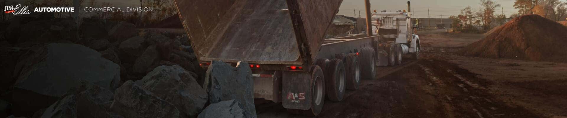 Rear three-quarter view of a semi-truck lifting an end-dump trailer to unload large rocks.