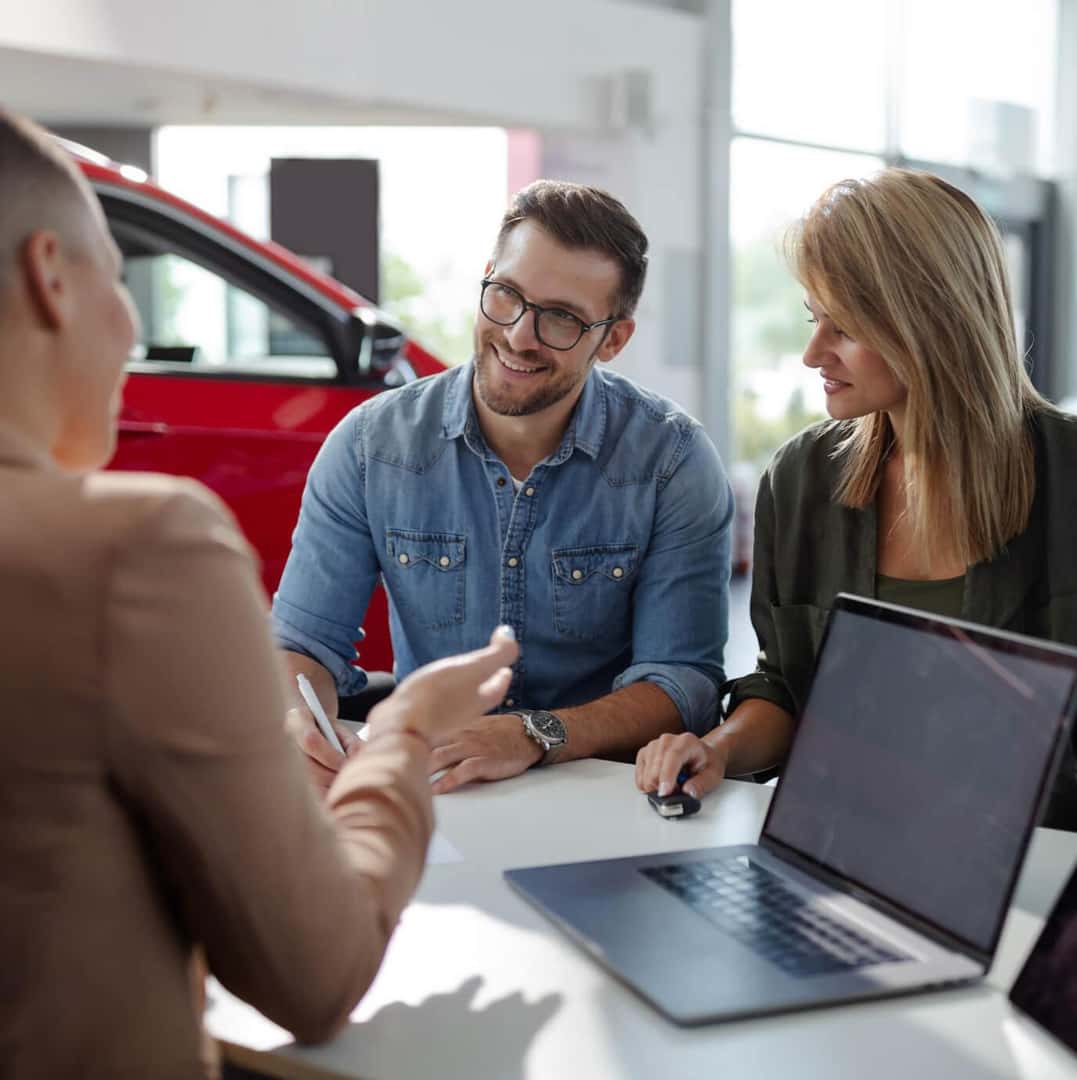 A financing agent talks to a smiling couple sitting at his desk.