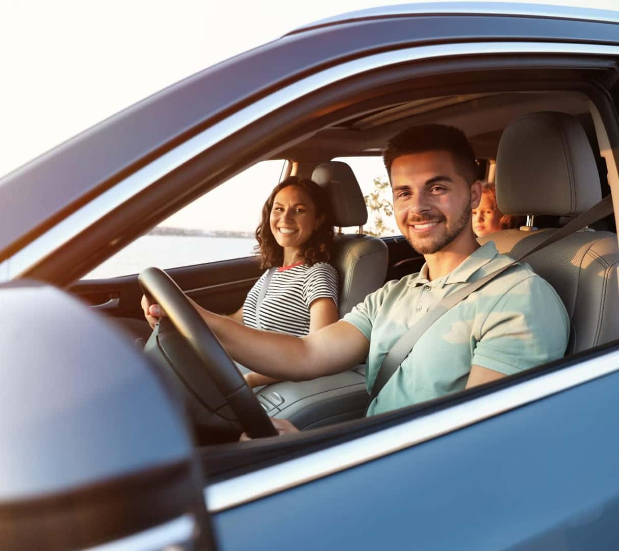 A smiling man drives a car with a woman in the front passenger seat and a child in the back. The sun sets outside, casting a warm glow inside.