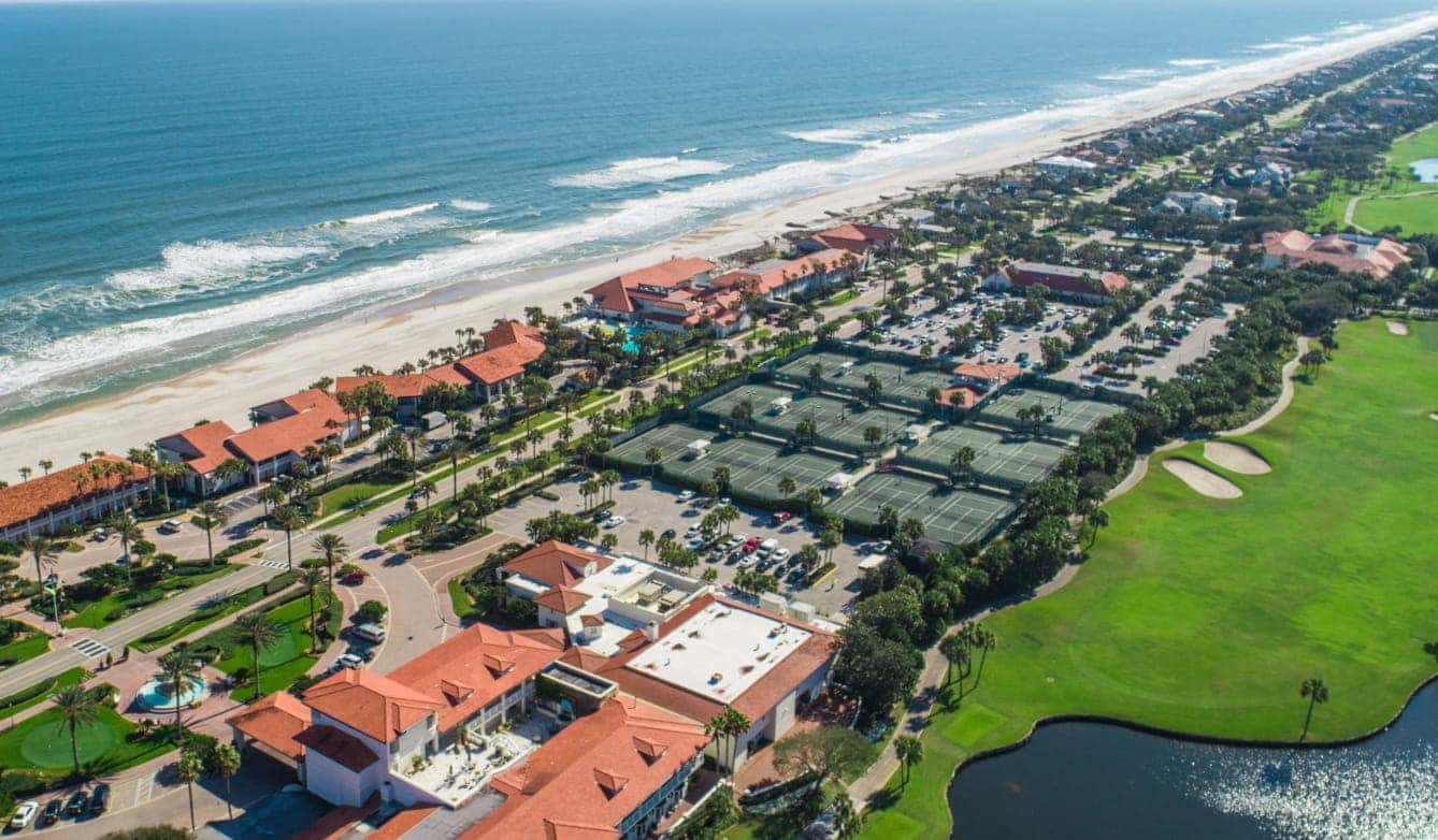 Aerial view of a beachfront resort with terracotta-roofed buildings, tennis courts, and lush greenery alongside the ocean.