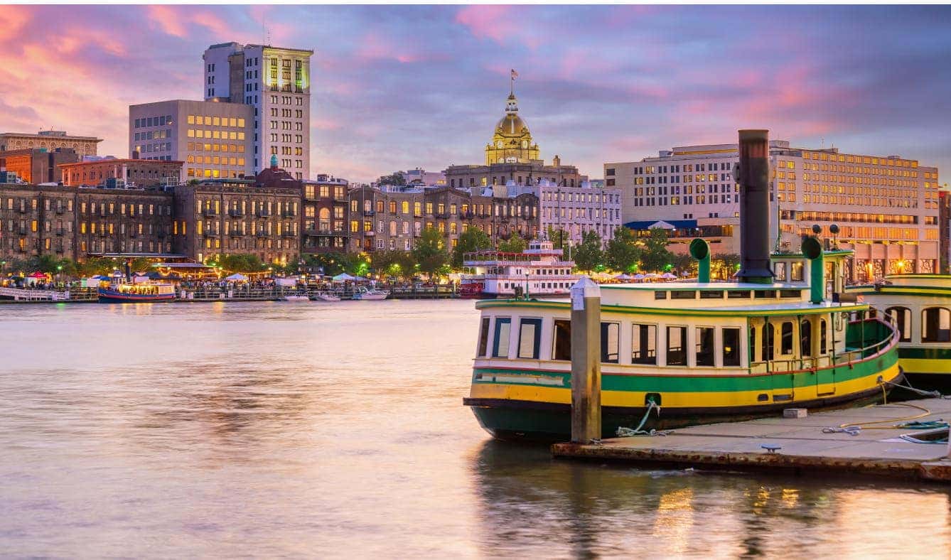 A serene riverside scene at dusk with a historic skyline, colorful sky, and two vintage-style boats docked in the foreground.