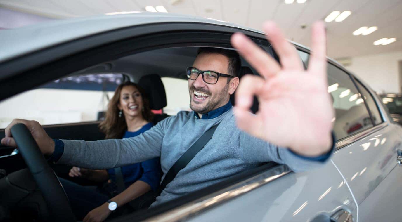 Smiling couple in a used car at a dealership, with the driver giving an OK gesture to show excellent value.