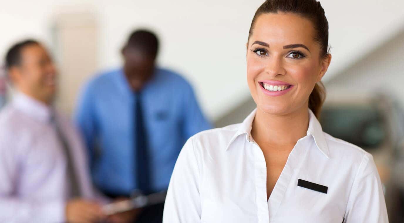 Smiling female dealership representative in a white shirt stands in a showroom with two customers blurred behind her.