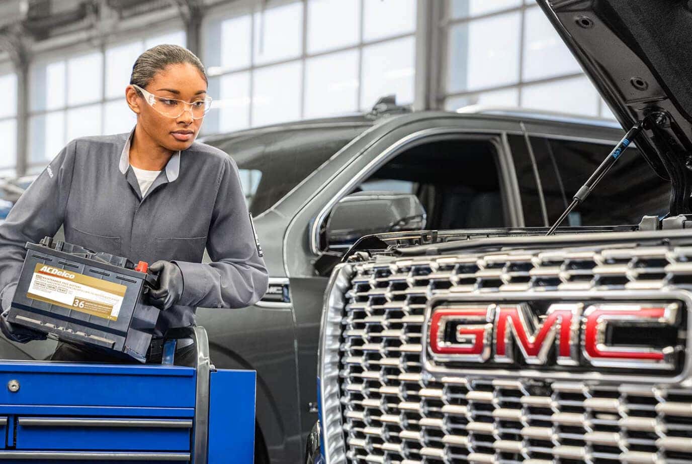 Certified female technician wearing safety glasses holding an ACDelco replacement battery for a GMC vehicle in a dealership service center.