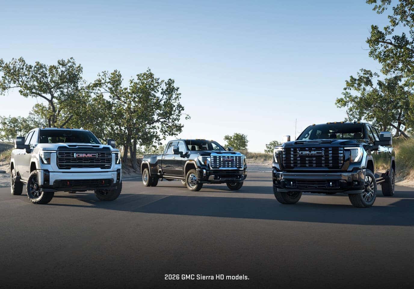 Front three-quarter lineup displaying three premium 2026 GMC Sierra HD trucks parked on an open paved stretch.