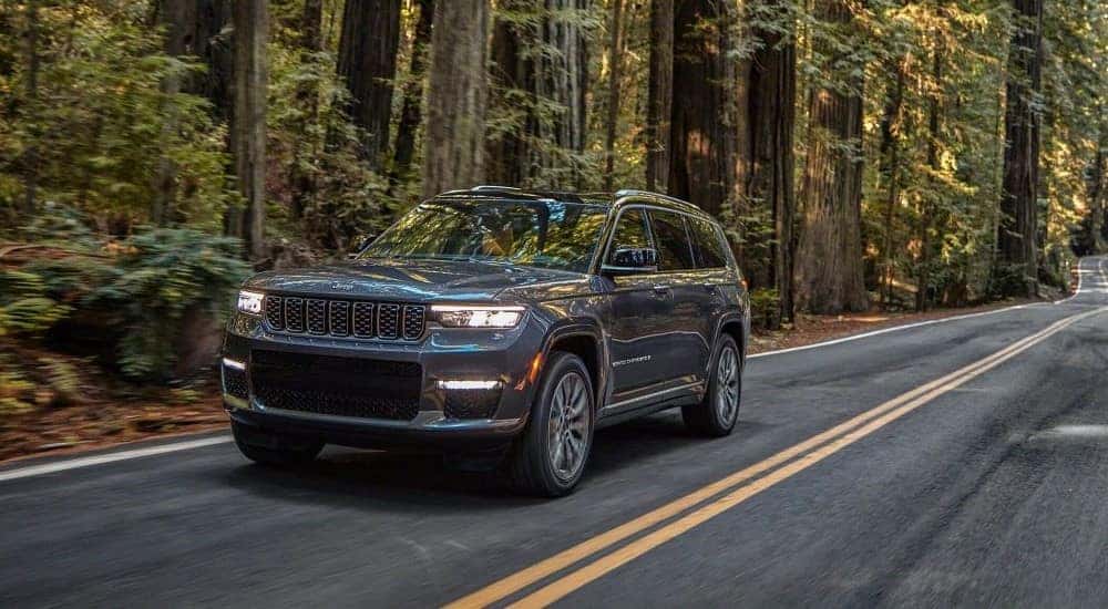 A dark grey 2021 Jeep Grand Cherokee L is shown driving on a tree-lined road.