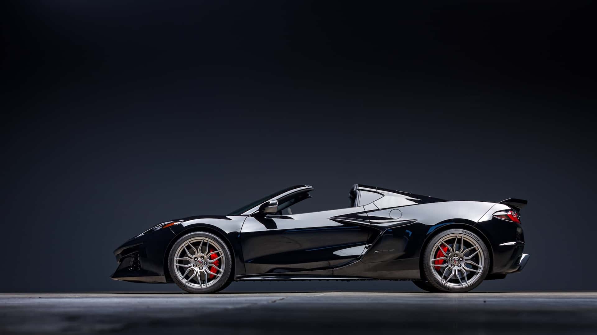 Side profile of a black Chevrolet Corvette Z06 convertible sports car parked in a studio setting with dramatic lighting, showcasing its sleek design, alloy wheels, and performance styling.