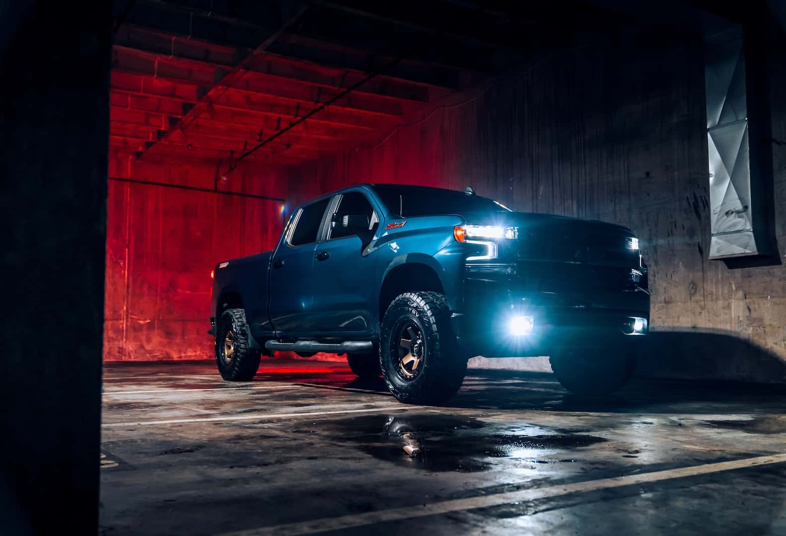 Chevrolet Silverado Z71 pickup truck parked in an industrial warehouse with red accent lighting, featuring off-road tires, illuminated headlights, and a bold, rugged appearance.