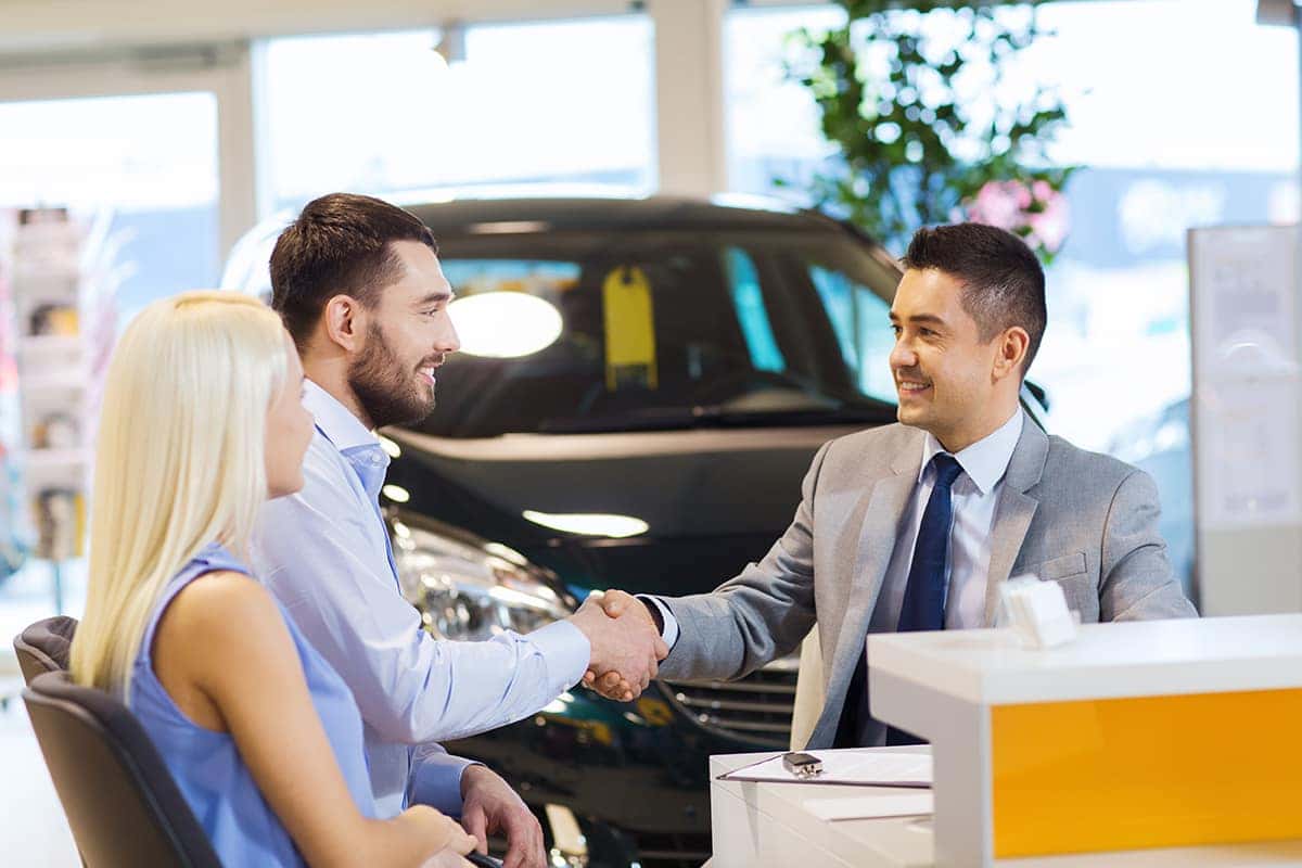 Customer and salesman shaking hands while happy lady is sitting in the driver's seat of car